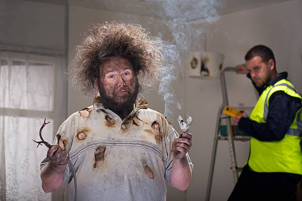 Image of a man holding two electrical cables. He's wearing a white t-shirt with burn-holes in them. His hair is fuzzy because of electrical shock. In the background, there's a ladder near the wall and an electrician wearing a hi-vis vest.