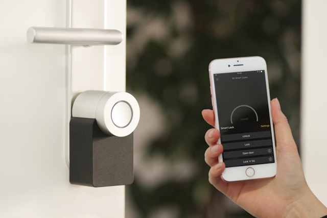 Image is a close-up of a white door with a handle. The door handle has a smart home lock on it. On the right-hand side, a female hand is holding an iPhone with a smart home security app open on the phone's screen.