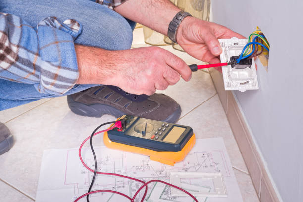Image is of an electrician working on an electrical socket in the wall. He has a meter with him.